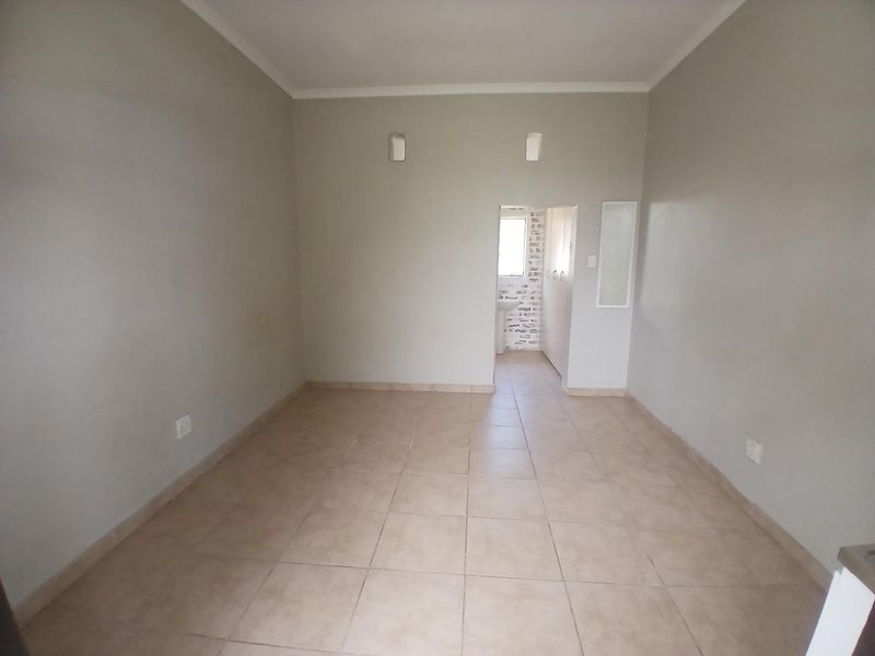 An empty, neutral-toned living area featuring tiled flooring, coving, and wall-mounted lighting, with a clear view into an adjacent room.