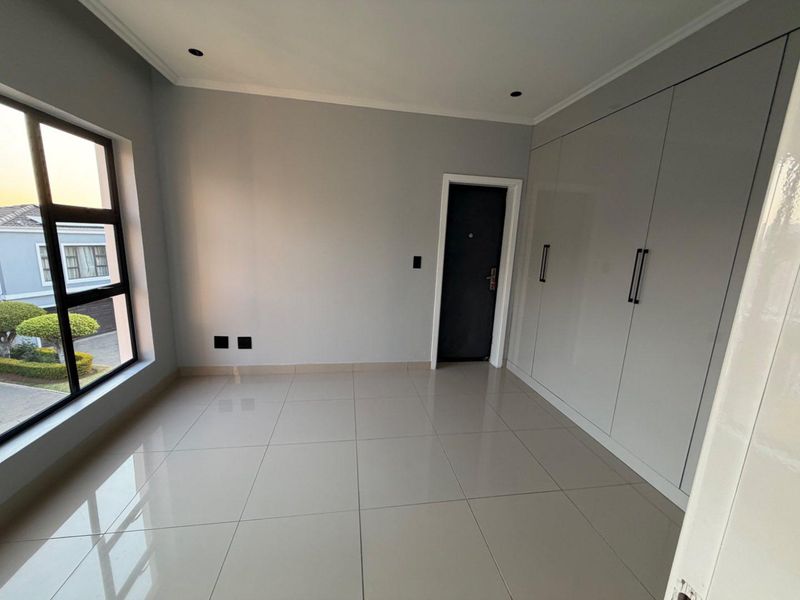 A bright, modern bedroom featuring neutral tiled flooring, built-in cupboards, and large windows providing natural light.