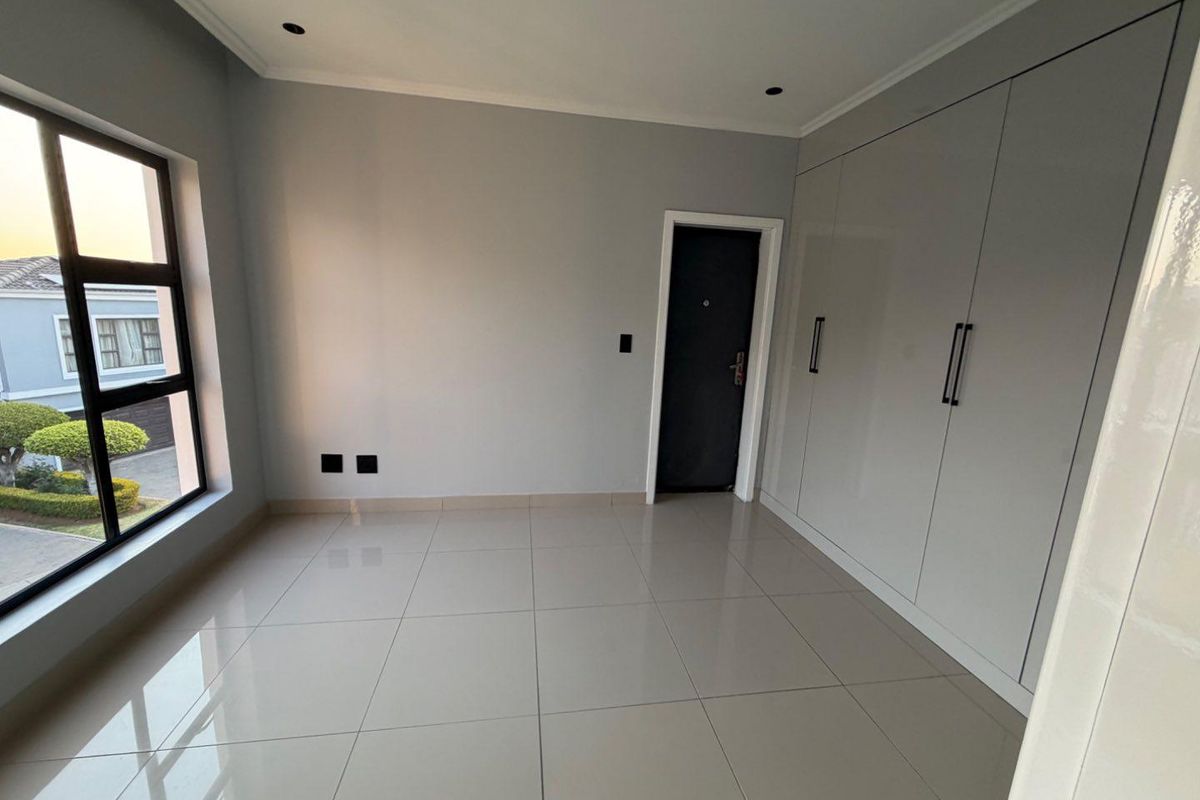 A bright, modern bedroom featuring neutral tiled flooring, built-in cupboards, and large windows providing natural light.