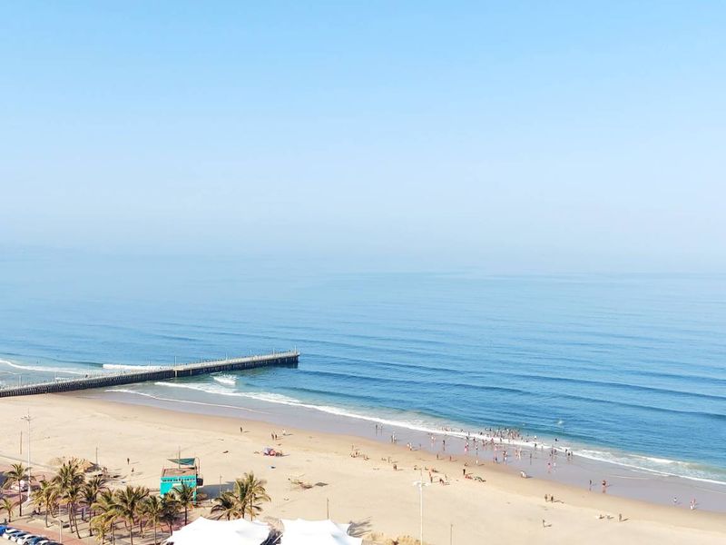 A panoramic elevated view of the coastline, featuring a sandy beach, ocean waves, and a pier.
