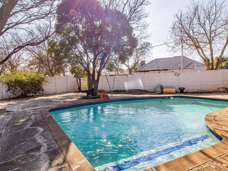 A backyard swimming pool area featuring stone paving and a perimeter boundary wall, set within a mature garden space.