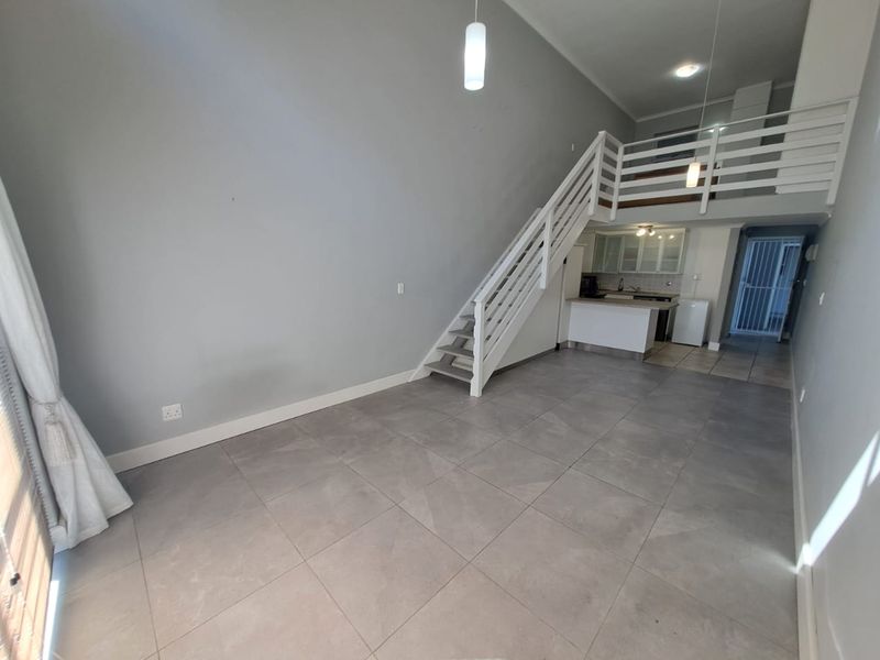 Spacious open-plan living area featuring large floor tiles, a staircase leading to a mezzanine level, and a compact kitchen area.
