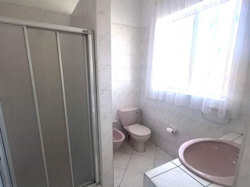 A dated bathroom featuring a shower enclosure, toilet, bidet, and a tiled vanity with a basin, finished with neutral floor and wall tiles.