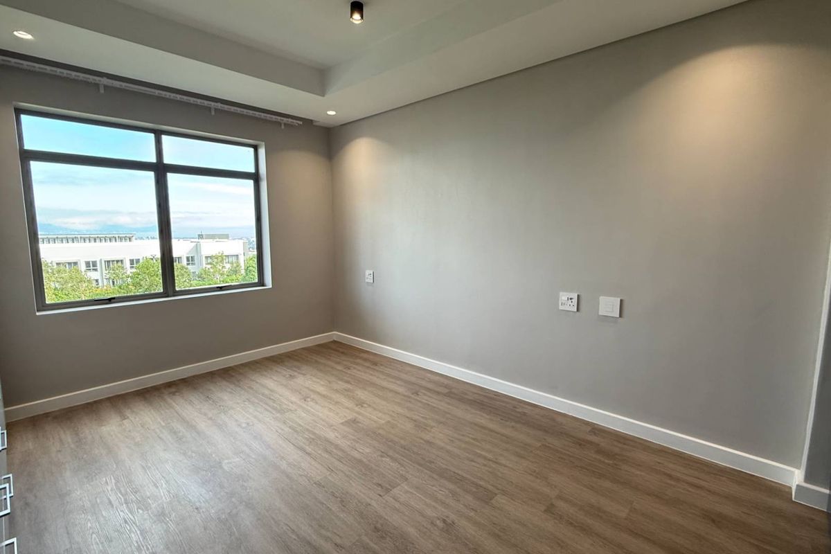 A bright, empty bedroom featuring modern laminate flooring, built-in cupboards, and a large window with a pleasant exterior view.