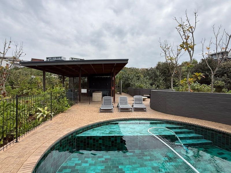 A well-maintained outdoor pool area featuring a tiled swimming pool, paved surround, and a covered patio structure set within a landscaped garden.