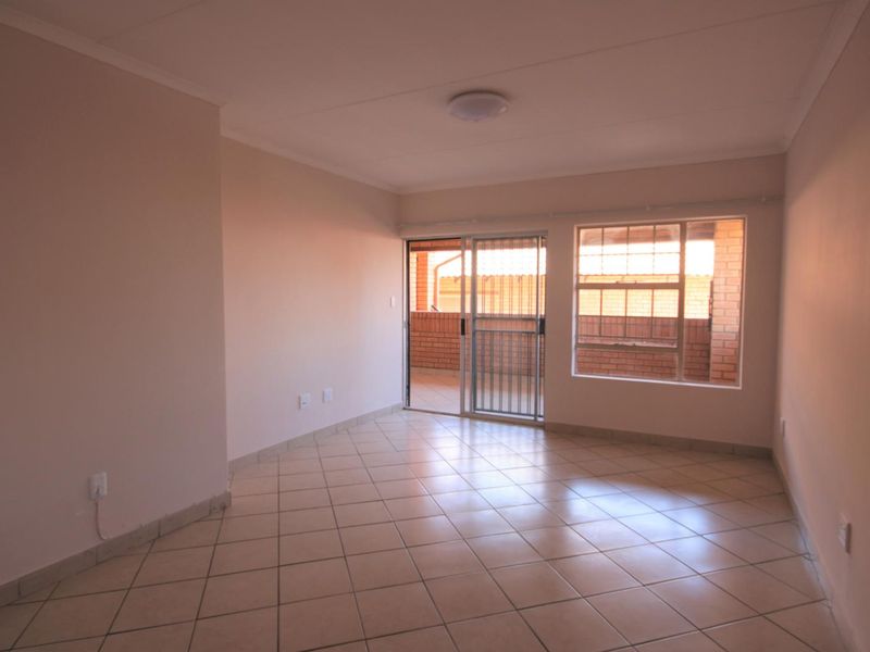 A bright, empty living room featuring neutral tiled flooring, a sliding glass door leading to a balcony, and large windows for natural light.