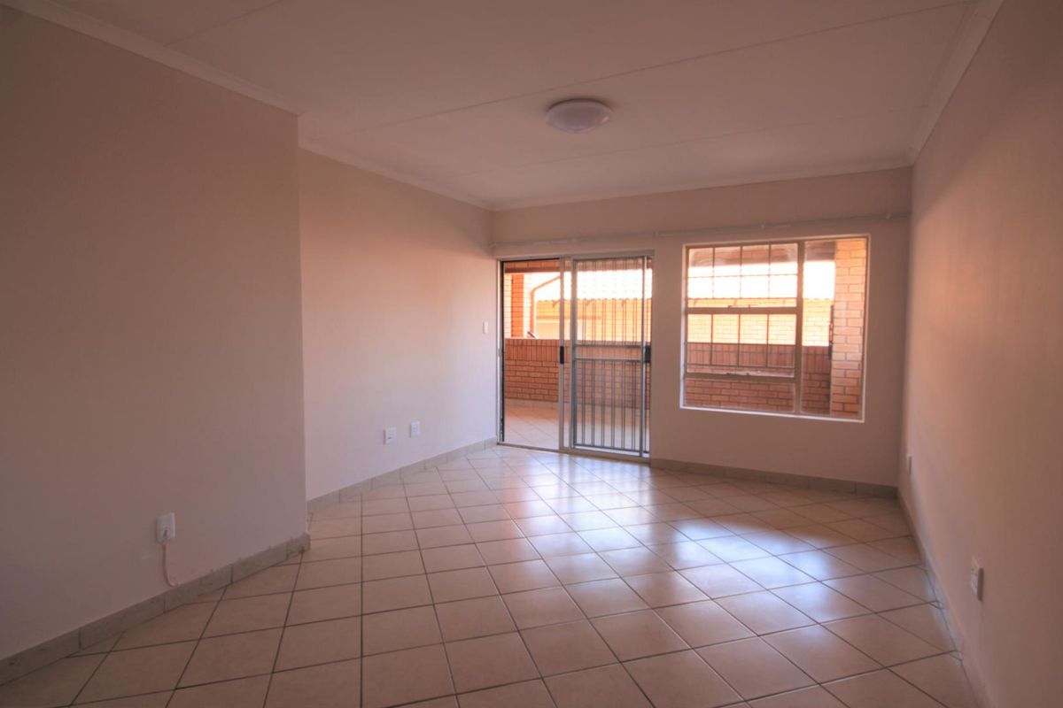A bright, empty living room featuring neutral tiled flooring, a sliding glass door leading to a balcony, and large windows for natural light.