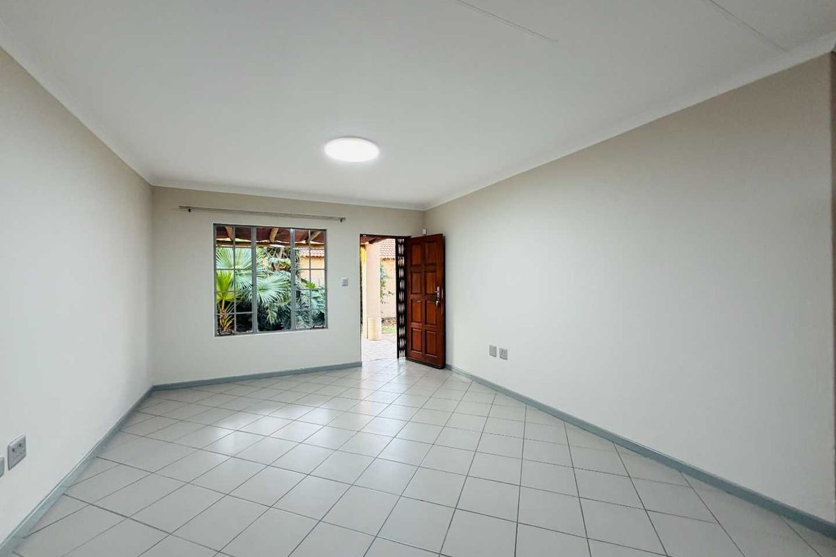 A bright, empty living room featuring neutral tiled flooring, a timber entrance door with security gate, and a window overlooking the garden.