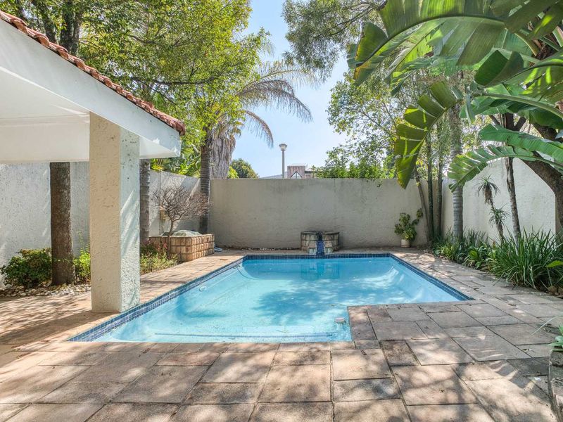 A private outdoor pool area featuring a tiled surround, a covered patio space, and a perimeter boundary wall with established garden greenery.