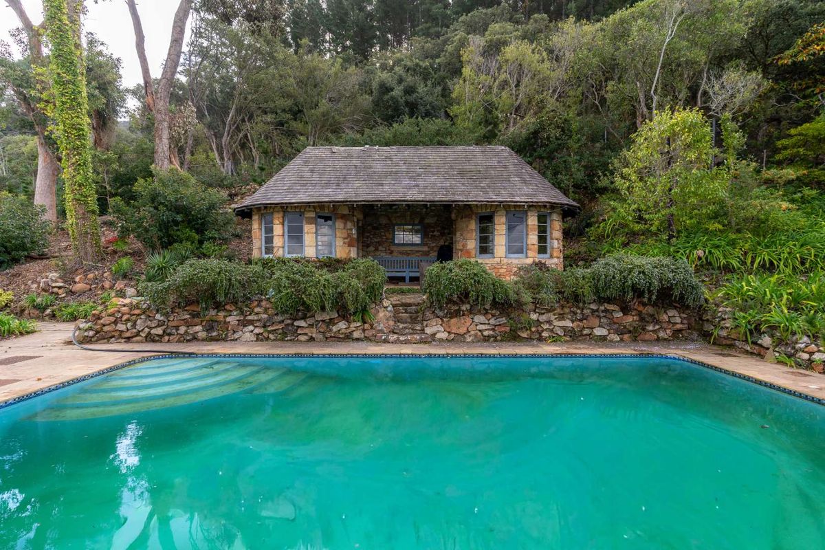 A rustic stone cottage overlooking a swimming pool, set against a lush, wooded hillside backdrop.