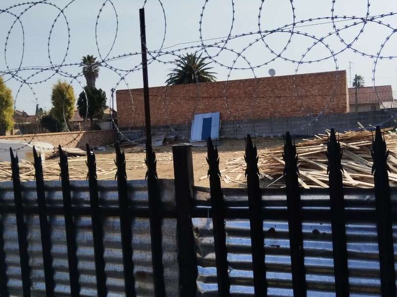 A vacant residential plot enclosed by metal fencing and topped with razor wire, featuring a brick boundary wall and scattered construction debris.