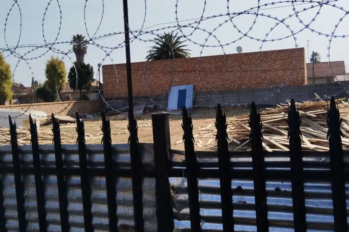 A vacant residential plot enclosed by metal fencing and topped with razor wire, featuring a brick boundary wall and scattered construction debris.