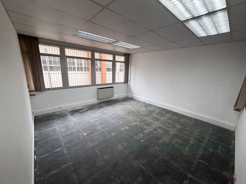 An empty office space featuring a suspended ceiling with recessed lighting, large windows, and a wall-mounted HVAC unit, ready for new flooring and finishes.