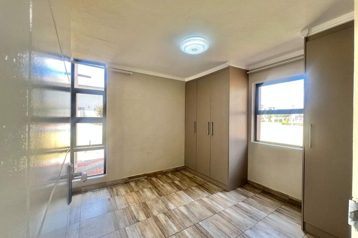 A bright, empty bedroom featuring modern wood-look tiled flooring, built-in cupboards, and large windows providing ample natural light.