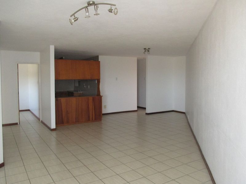 Spacious open-plan living area featuring neutral ceramic floor tiles, built-in kitchen cabinetry, and modern track lighting in a clean, vacant setting.