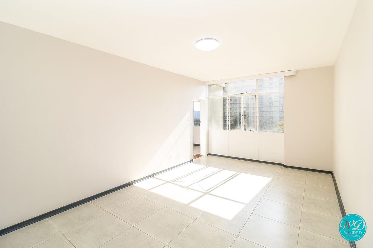A bright, empty living room featuring neutral wall tones, tiled flooring, and a large window allowing for ample natural light.