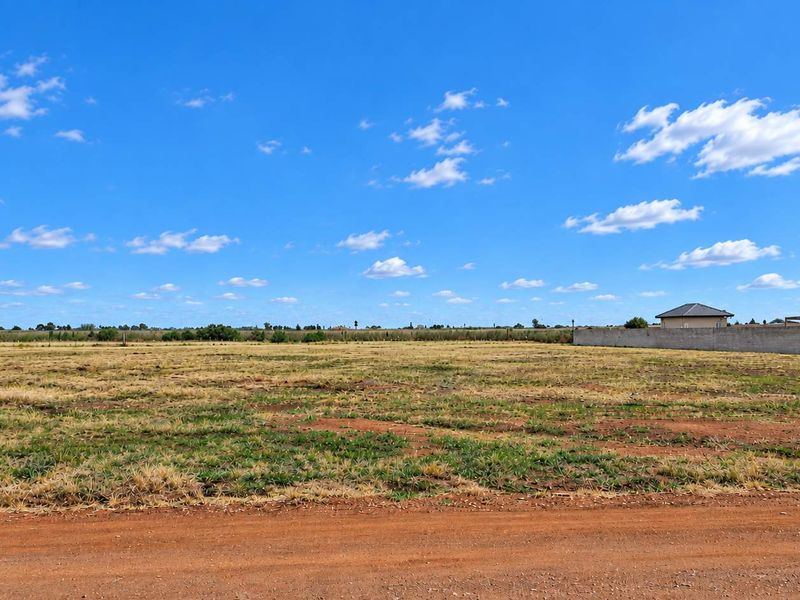 A wide, level vacant residential plot featuring natural grass cover, bordered by a perimeter wall with adjacent residential development visible.