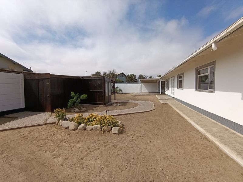 Rear exterior view featuring a paved walkway, sandy garden area, and a timber structure alongside the main house building.