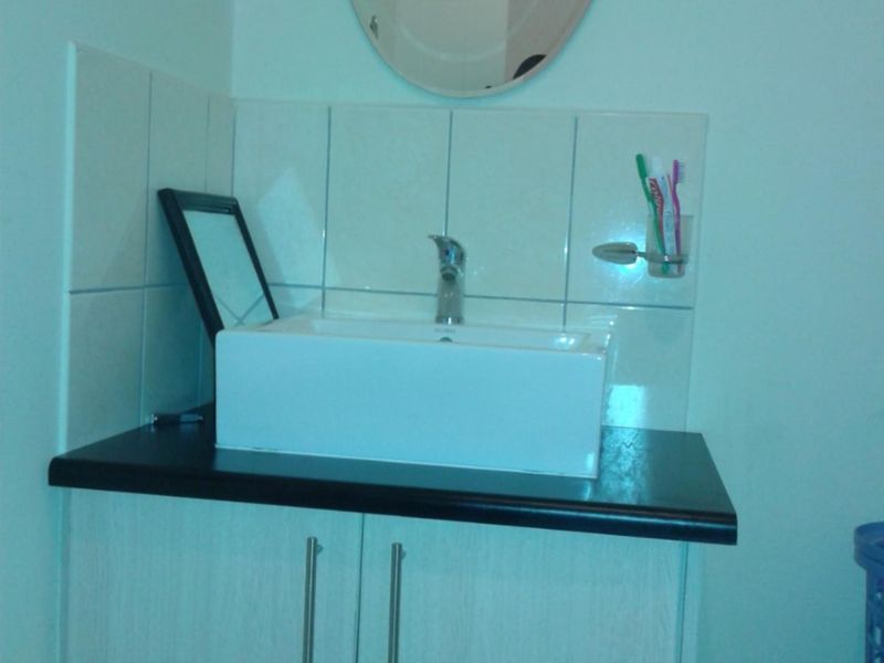 A bathroom vanity featuring a modern vessel sink, dark countertop, and light wood-finish cabinetry with a wall-mounted mirror and tiled splashback.