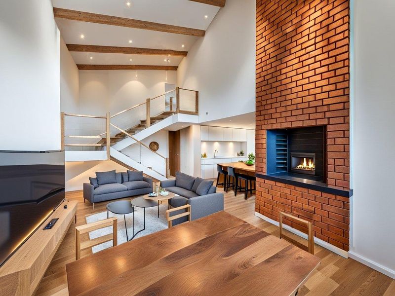 Spacious open-plan living area featuring a double-volume ceiling with exposed timber beams, a striking brick fireplace, and a modern staircase.