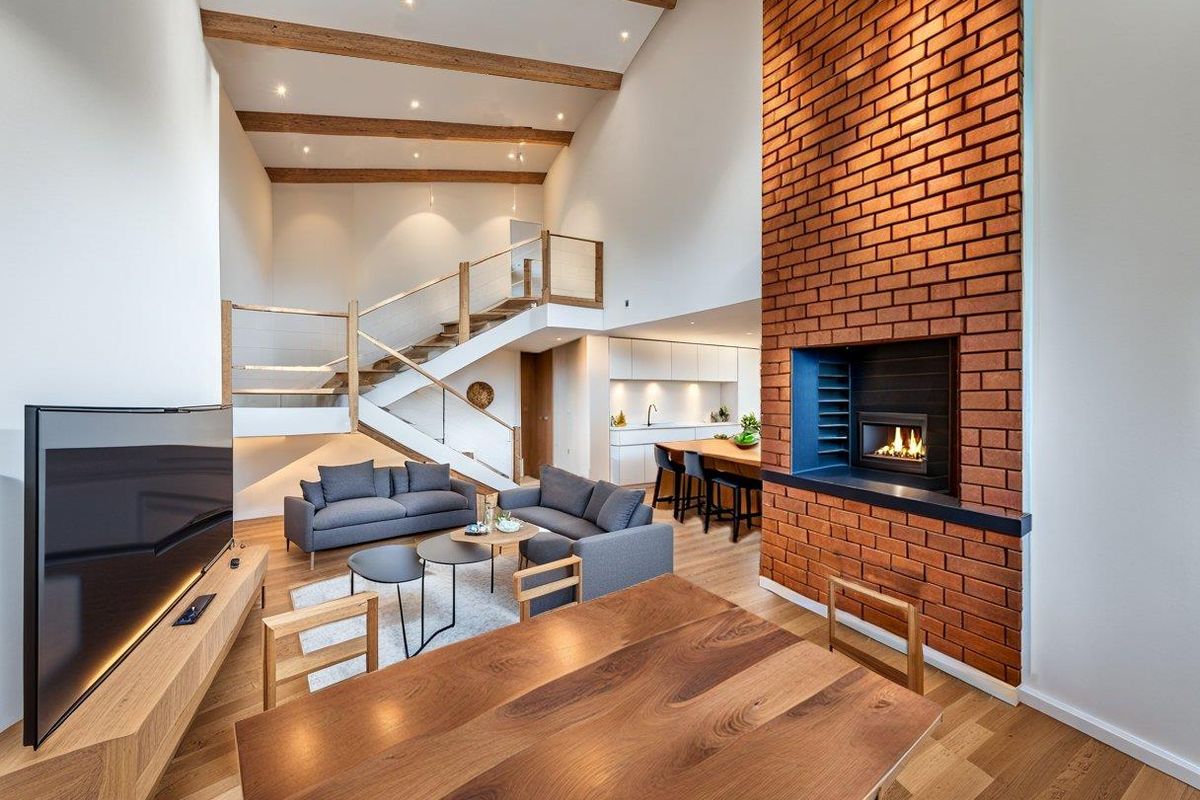 Spacious open-plan living area featuring a double-volume ceiling with exposed timber beams, a striking brick fireplace, and a modern staircase.