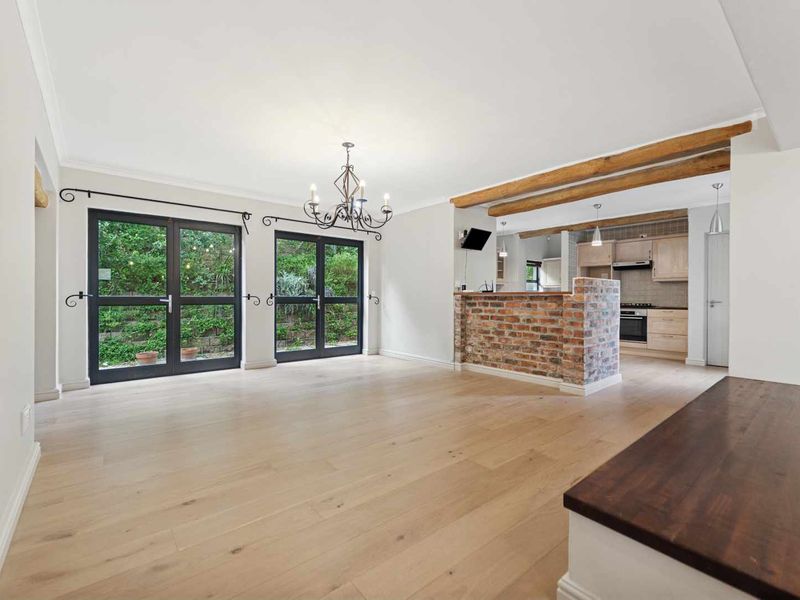 Spacious open-plan living and kitchen area featuring light timber flooring, exposed brick breakfast bar, timber ceiling beams, and double french doors leading to the garden.