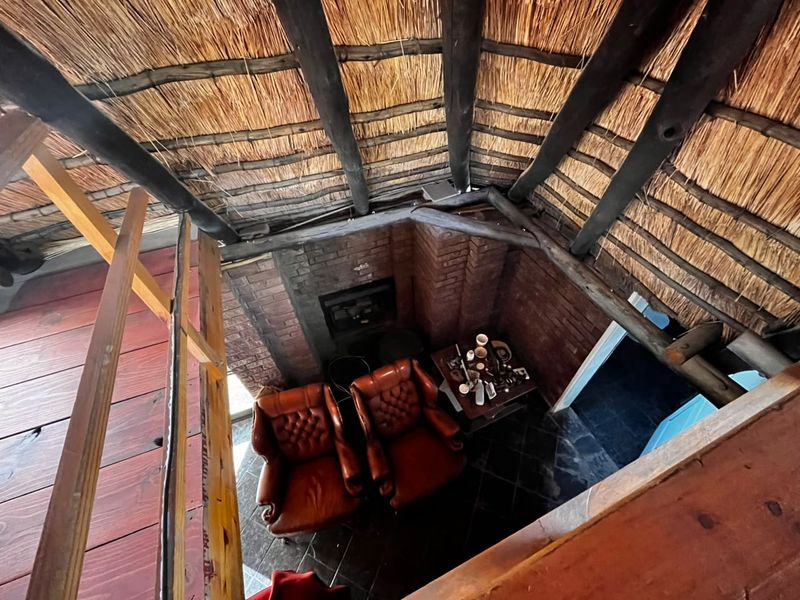 A unique living area featuring a high-pitched thatched roof with exposed timber rafters, a brick fireplace, and slate flooring, viewed from a wooden mezzanine level.