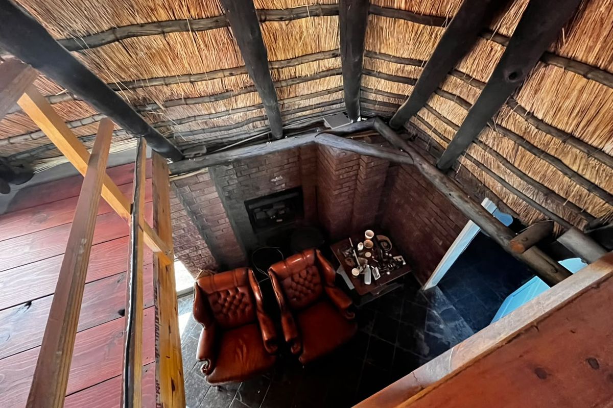 A unique living area featuring a high-pitched thatched roof with exposed timber rafters, a brick fireplace, and slate flooring, viewed from a wooden mezzanine level.