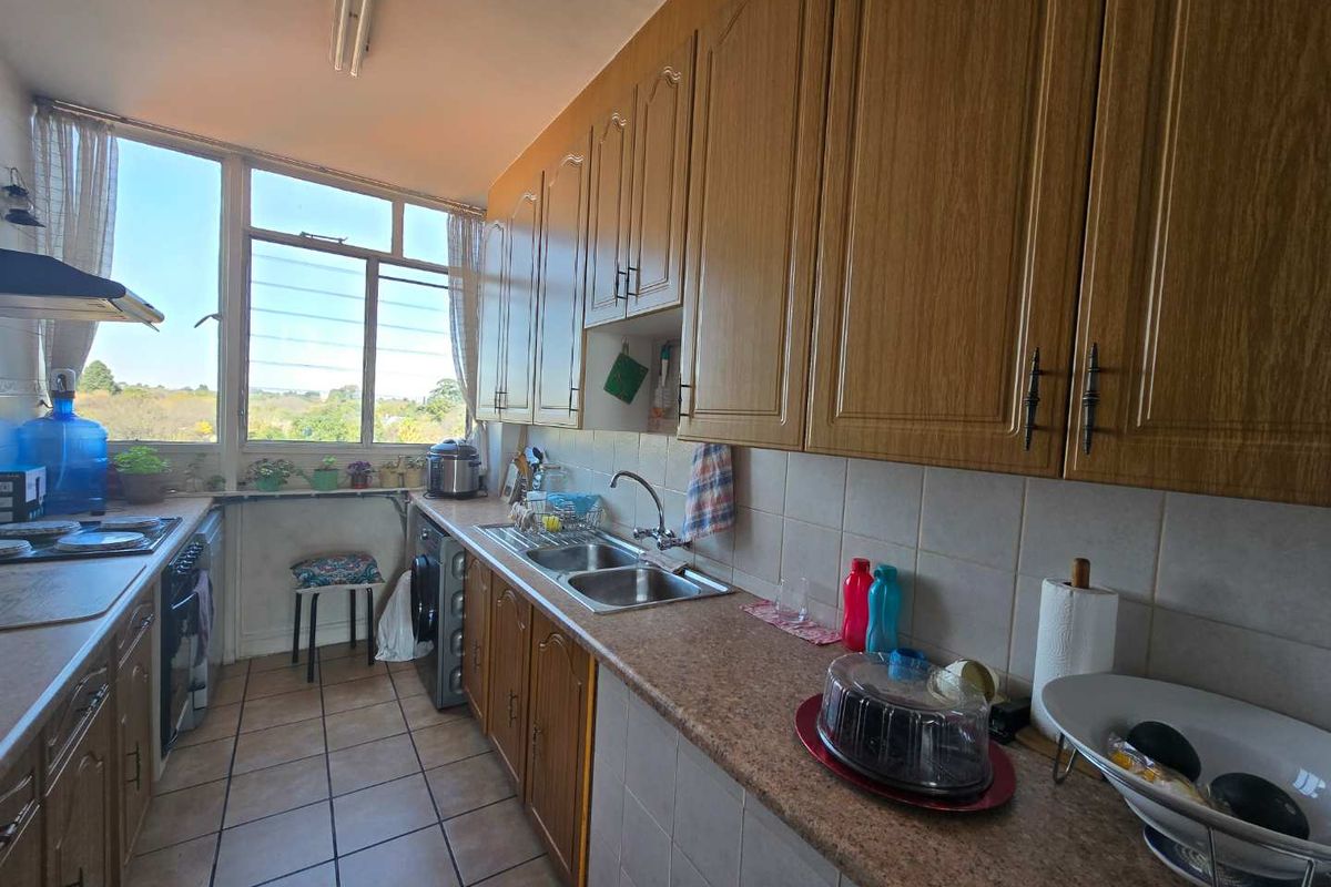 A functional galley-style kitchen featuring wood-look cabinetry, laminate countertops, a double basin sink, and tiled finishes with natural light.