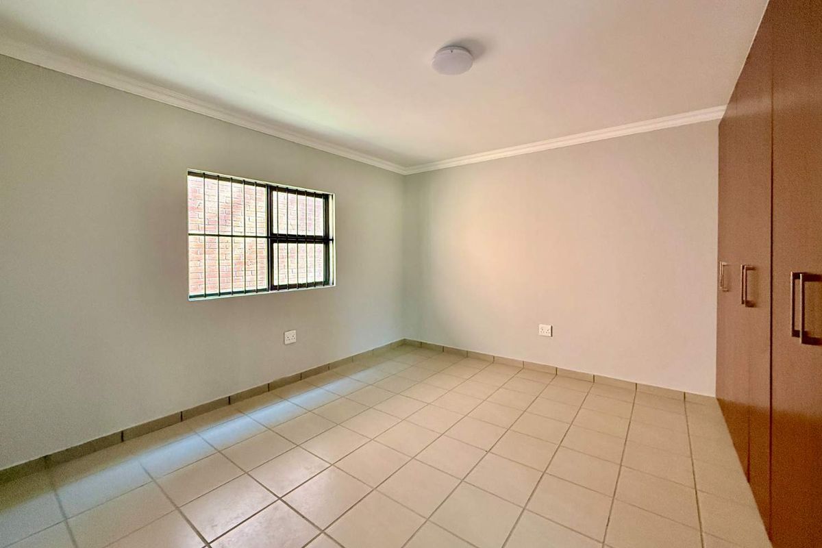 A clean, empty bedroom featuring neutral tiled flooring, built-in wooden cupboards, and a window with security bars.