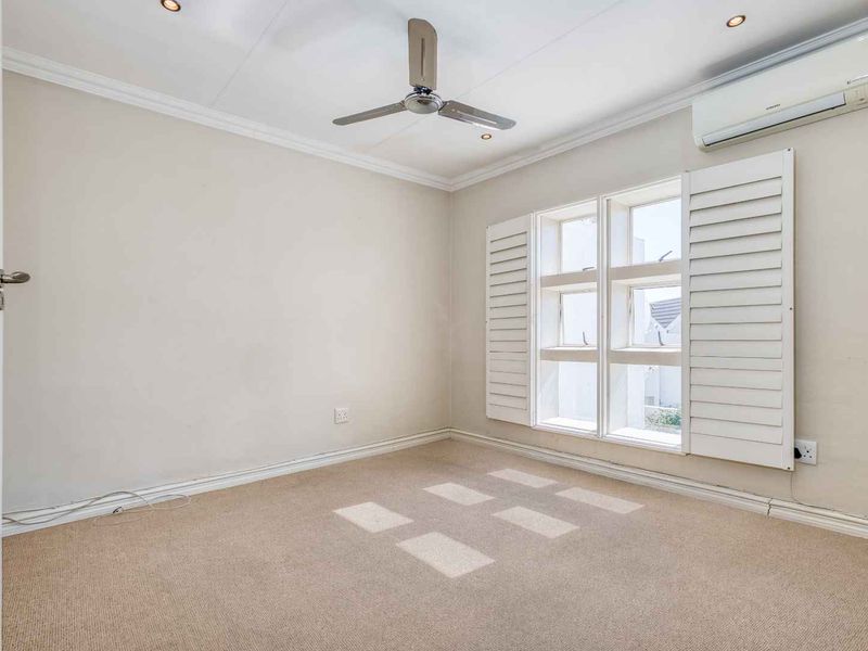 A bright, empty bedroom featuring neutral carpeting, a ceiling fan, split-system air conditioning, and white plantation shutters.