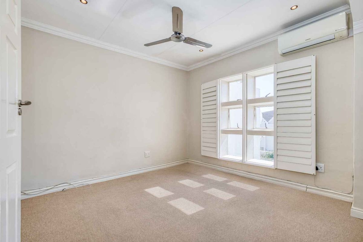 A bright, empty bedroom featuring neutral carpeting, a ceiling fan, split-system air conditioning, and white plantation shutters.