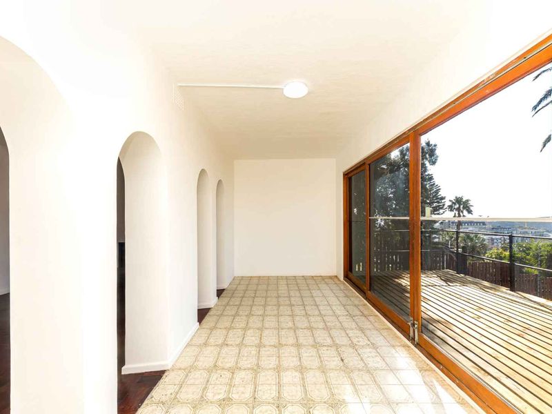 A bright, empty covered balcony featuring arched architectural details, tiled flooring, and large sliding glass doors opening onto a timber deck with outdoor views.