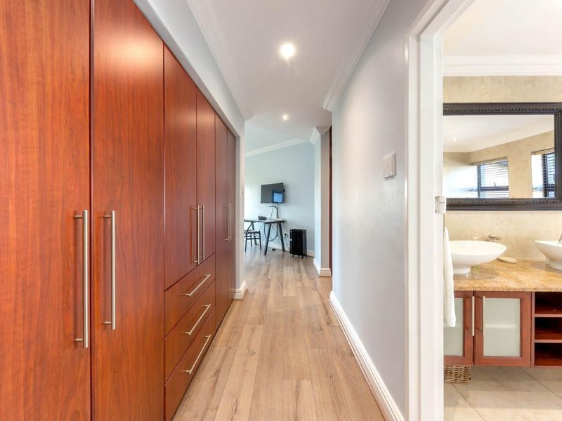 A well-lit hallway featuring extensive built-in wooden cabinetry, laminate flooring, and a view into an adjacent bathroom with dual vessel sinks.