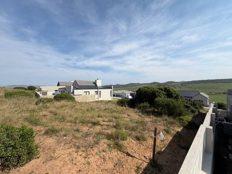 A view of a vacant residential plot featuring natural fynbos vegetation and a concrete boundary wall, overlooking neighboring properties and distant hills.