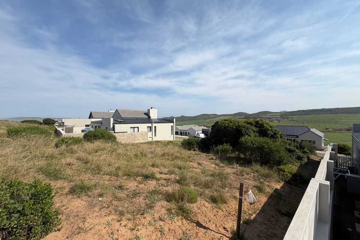 A view of a vacant residential plot featuring natural fynbos vegetation and a concrete boundary wall, overlooking neighboring properties and distant hills.