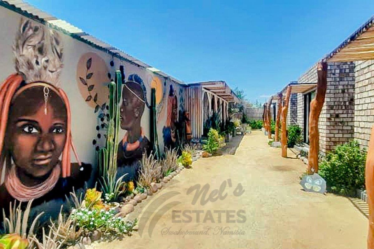 A landscaped outdoor walkway featuring a decorative mural wall, natural wood support posts, and a sandy path leading between guest house units.