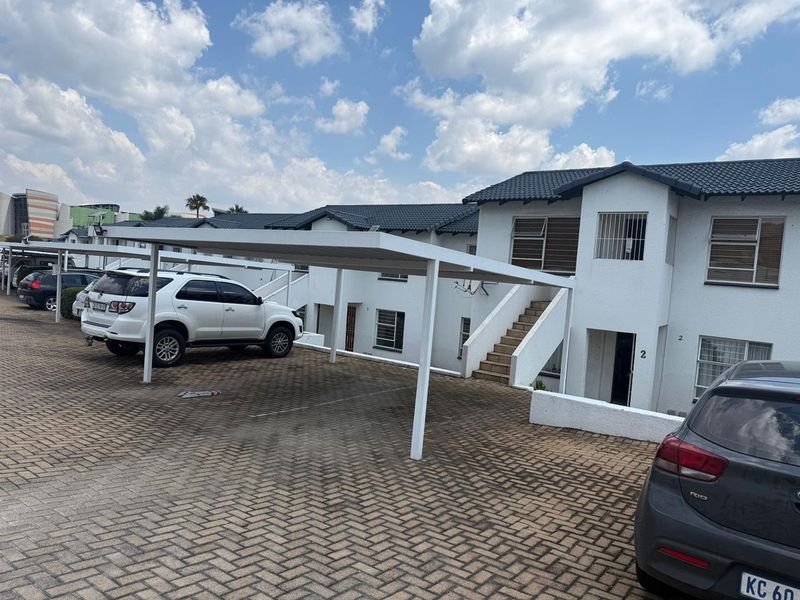 Front exterior view of a townhouse complex featuring a paved driveway, covered carports, and white facade with security bars on windows.