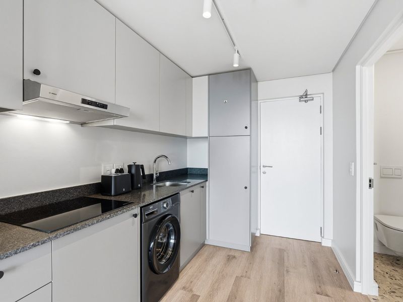 Modern galley kitchen featuring sleek grey cabinetry, stone countertops, integrated appliances, and high-quality laminate flooring, with adjacent access to a bathroom.