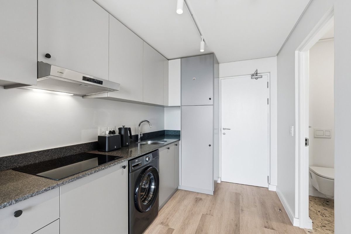 Modern galley kitchen featuring sleek grey cabinetry, stone countertops, integrated appliances, and high-quality laminate flooring, with adjacent access to a bathroom.