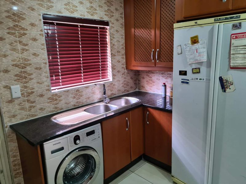 A functional kitchen area featuring dark granite countertops, wooden cabinetry, a double bowl sink, and tiled backsplash with integrated laundry plumbing.