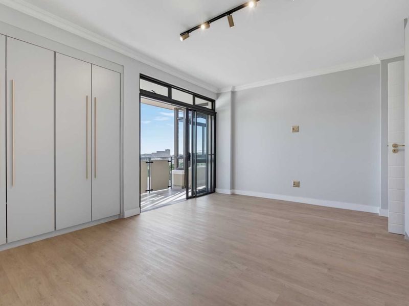 A bright, modern bedroom featuring built-in cupboards, laminate flooring, and direct access to a private balcony through sliding glass doors.