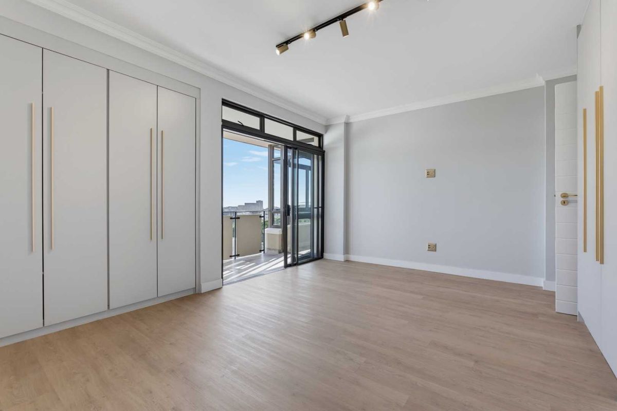 A bright, modern bedroom featuring built-in cupboards, laminate flooring, and direct access to a private balcony through sliding glass doors.