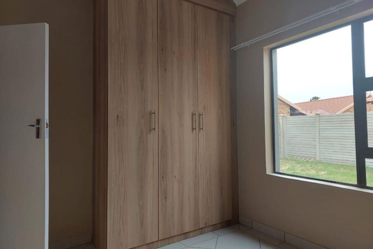 A bedroom featuring built-in wooden cupboards, tiled flooring, and a large window providing natural light and a view of the rear garden area.
