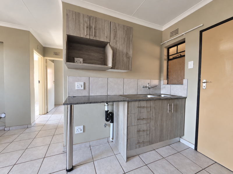 A compact kitchen area featuring wood-look cabinetry, granite countertops, a stainless steel sink, and neutral tiled flooring.