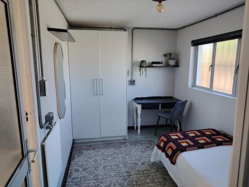 A compact bedroom featuring a built-in wardrobe, exposed electrical conduit, and an aluminium-framed window, providing a functional and simple living space.