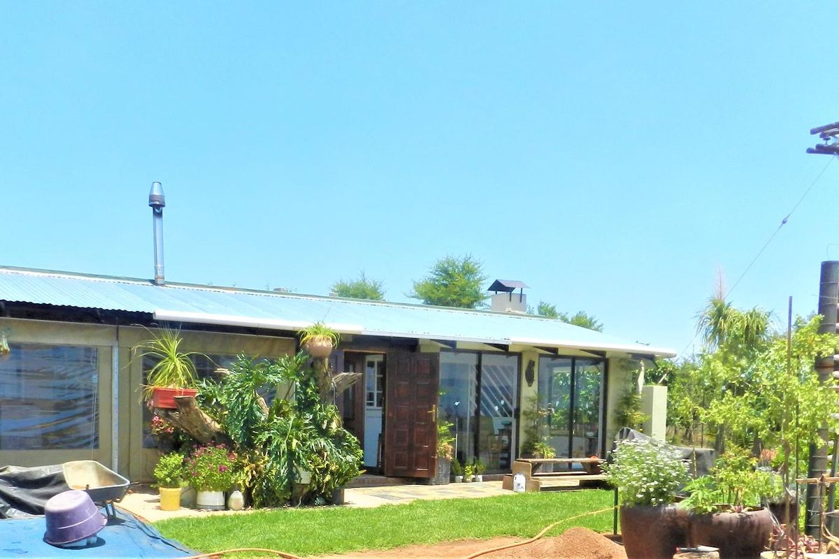 Rear view of the farm dwelling featuring a corrugated metal roof, sliding glass doors, and a paved patio area set within a garden.