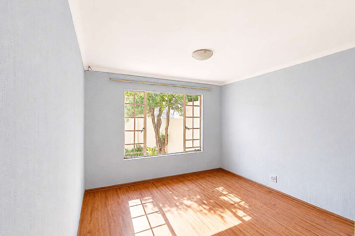 A bright, empty bedroom featuring laminate flooring, textured walls, and a large steel-framed window overlooking the garden.
