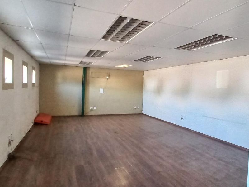 A spacious, open-plan industrial office or utility area featuring wood-look laminate flooring, recessed ceiling lighting, and multiple small square windows.