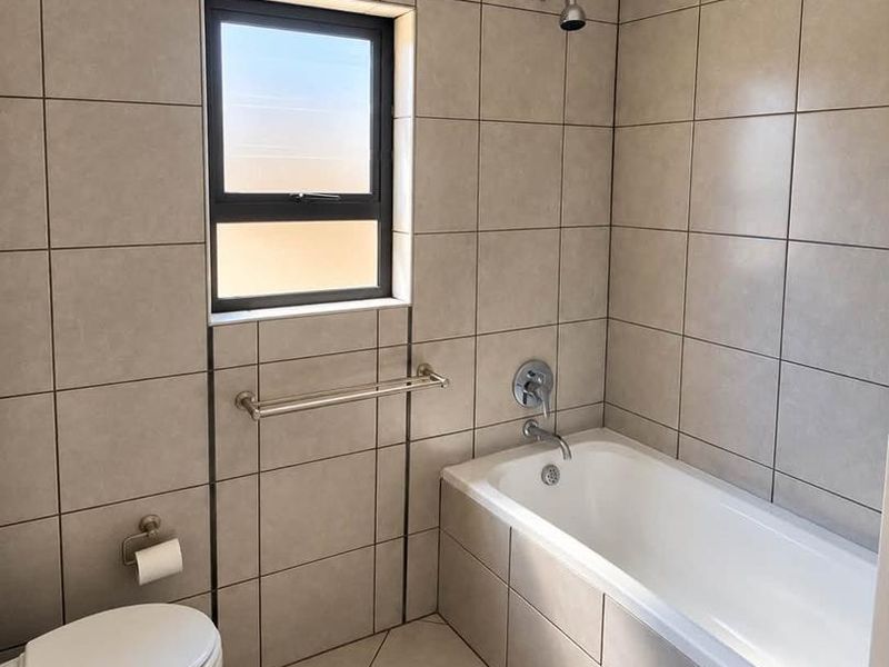 A clean, modern bathroom featuring neutral wall and floor tiles, a built-in bathtub with shower head, and standard sanitary fixtures.